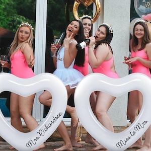 Puede incluir: Dos accesorios de cabina de fotos inflables en forma de corazón blanco con el texto "Mrs. Richardson" y "Miss to Mrs." impreso en ellos. Un grupo de mujeres posan con los accesorios, vistiendo vestidos rosas y blancos y sosteniendo copas de vino.