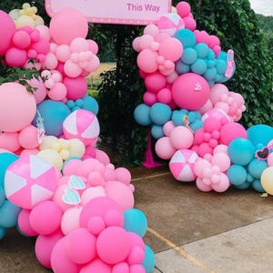 May include: A balloon arch made of pink, blue and white balloons. The arch is decorated with beach ball balloons and a sign that says "This Way".