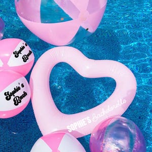 Puede incluir: Flotador de piscina en forma de corazón rosa con el texto "Sophie's Bachelorette" impreso en él. También hay varios balones de playa rosas y blancos en la piscina.