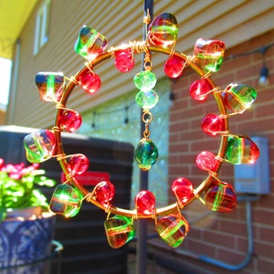 May include: A gold wire circle with red and green glass beads hanging from it. The beads are iridescent and have a striped pattern. The circle is hanging from a hook in a brick wall.