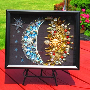 May include: A framed artwork depicting a crescent moon and sun made of colorful jewels and rhinestones. The moon is blue and white, while the sun is gold and red. The artwork is displayed on a black easel.