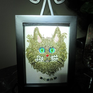 May include: A framed artwork of a cat's face made from gold and green beads. The cat has large green eyes, a red nose, and a black and white nose.
