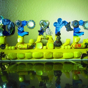 May include: A yellow wooden sign with the word "PEACE" spelled out in blue and white mosaic tiles. The sign is decorated with various small trinkets, including butterflies, hearts, and beads. The sign is reflecting in a dark surface.