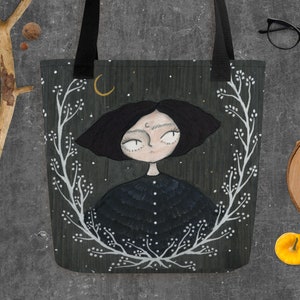 May include: A black tote bag featuring a white floral design and a black and white illustration of a girl with a crescent moon above her head. The background is a dark green with white stars.