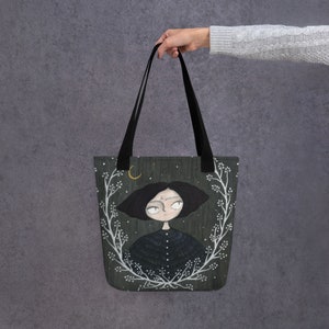 May include: A black tote bag with a dark green background featuring a white floral wreath and a cartoon illustration of a girl with black hair and a crescent moon. The girl is wearing a black dress with white dots.