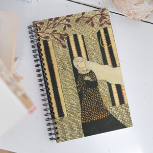 May include: A spiral bound notebook with a colorful illustration of a woman with long white hair standing in a stone archway. The woman is wearing a black dress with a gold and orange pattern. The background is a stone wall with a crescent moon in the upper right corner.