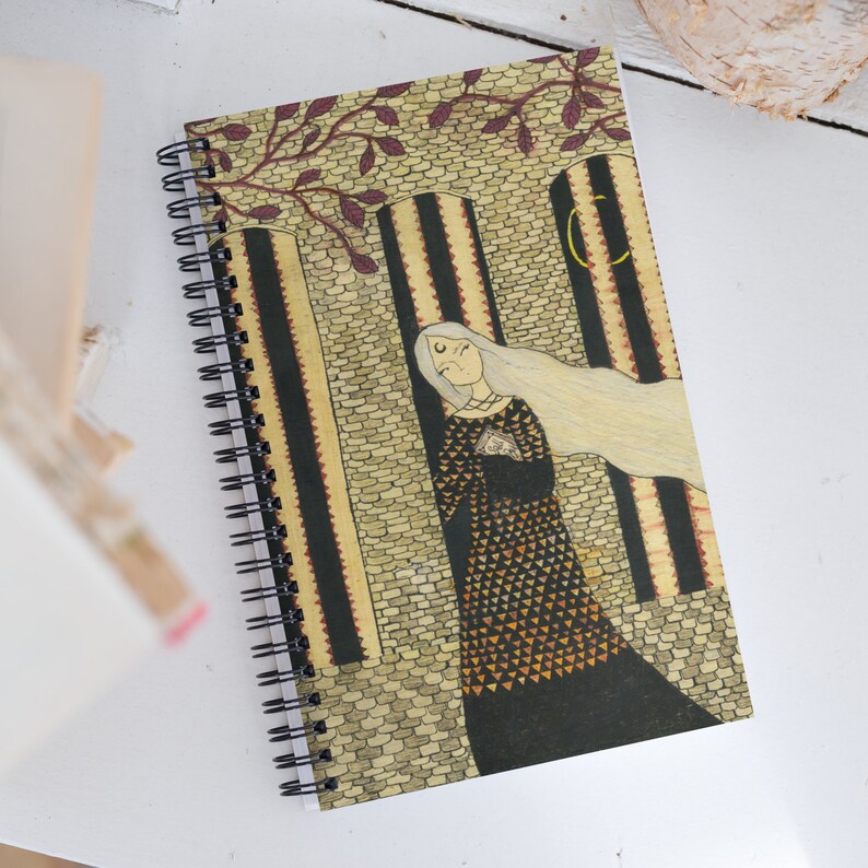 May include: A spiral bound notebook with a colorful illustration of a woman with long white hair standing in a stone archway. The woman is wearing a black dress with a gold and orange pattern. The background is a stone wall with a crescent moon in the upper right corner.