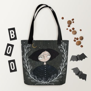 May include: A black tote bag featuring a white floral design and a black and white illustration of a girl with a crescent moon above her head. The background is a dark green with white stars.