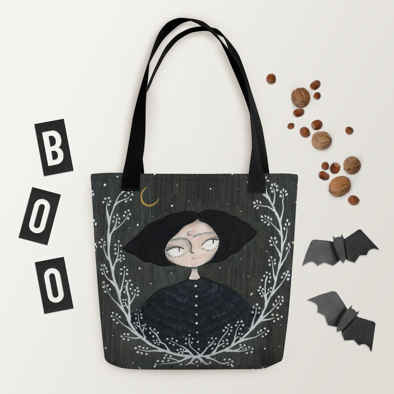 May include: A black tote bag featuring a white floral design and a black and white illustration of a girl with a crescent moon above her head. The background is a dark green with white stars.