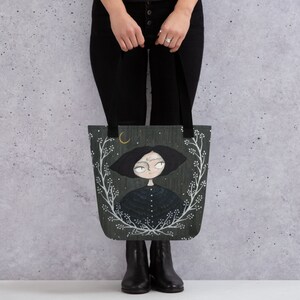 May include: A dark green tote bag featuring a white floral design and a black and white illustration of a girl with a crescent moon above her head.