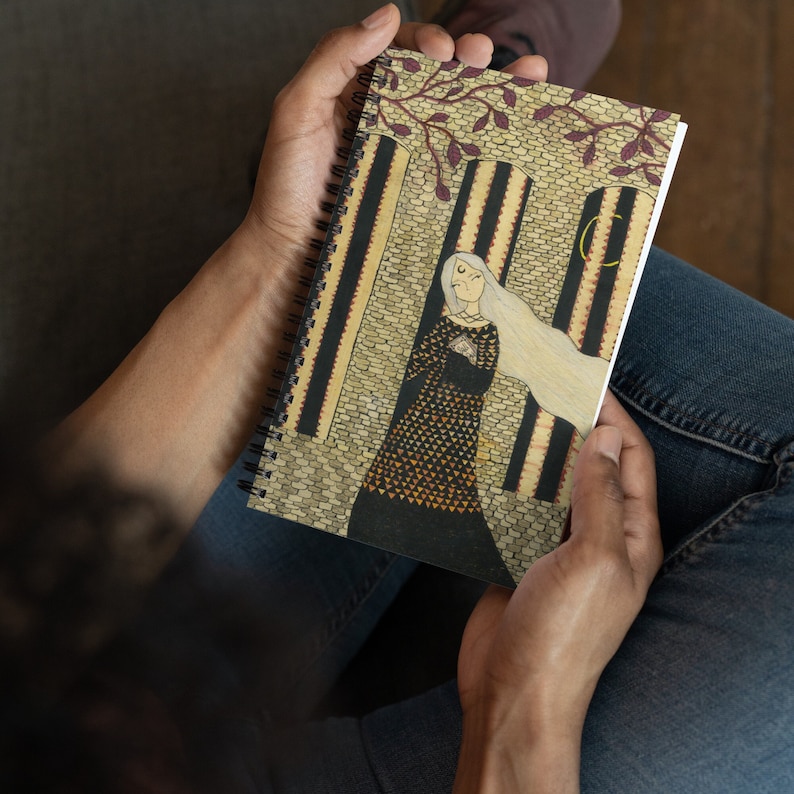 May include: A spiral bound notebook with a beige cover featuring an illustration of a woman in a black and gold dress standing in a doorway. The woman has long white hair and is holding a small object in her hands. The background is a brick wall with a window and a crescent moon.
