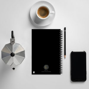 May include: A black spiral bound notebook with a gold crescent moon logo and the text "Mooncraft Instagram: Mooncraftdesigns". The notebook is on a white surface with a silver coffee pot, a cup of coffee, a pencil, and a black smartphone.