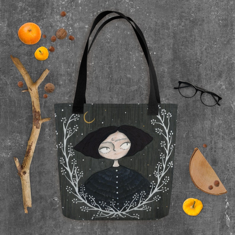 May include: A black tote bag featuring a white floral design and a black and white illustration of a girl with a crescent moon above her head. The background is a dark green with white stars.