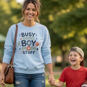 May include: A woman and a young boy are walking on grass. The woman wears a light blue sweatshirt with the text "Busy Doing Boy Mom Stuff". The boy holds a football, wearing a red t-shirt and khaki shorts. They are in a park.