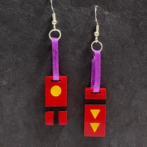 May include: A pair of red earrings with silver hooks and purple ribbon accents. One earring features a yellow circle, the other has two yellow downward-pointing triangles. The earrings are on a dark background.
