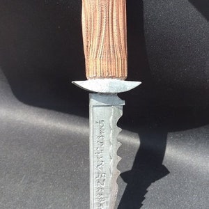 May include: A decorative dagger with a textured, light brown handle and a silver blade. The blade has a serrated edge and etched text. The handle is a light brown color, and the blade is silver.