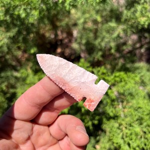 Puede incluir: Una punta de flecha rosa pálido y roja, de forma triangular y con base dentada, sostenida en la mano. La punta de flecha es de piedra y tiene una punta afilada. El fondo es una vista borrosa de follaje verde.