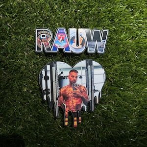 May include: A black and white heart-shaped sticker with a dripping effect. The sticker features a photo of a man with tattoos, wearing a black tank top, and holding a phone. The text "RAUW" is printed in blue and white on a separate sticker.