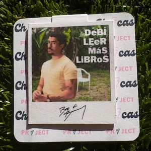 May include: A white square card with a photo of a person wearing a yellow shirt and a black watch. The photo is surrounded by a black border and the text "DeBi LEER Más LIBROS" is printed in black on the card. The card has a pink heart and the text "PROJECT" printed on the bottom.