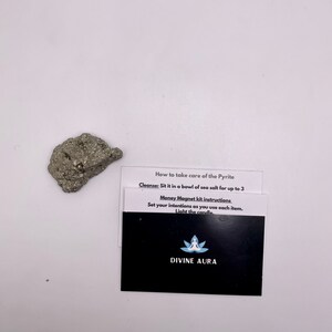 May include: A small, gray, metallic rock with a rough texture. A black card with white text reads "How to take care of the Pyrite Cleanse: Sit it in a bowl of sea salt for up to 3 Money Magnet kit instructions Set your intentions as you use each item. Light the candle. DIVINE AURA"