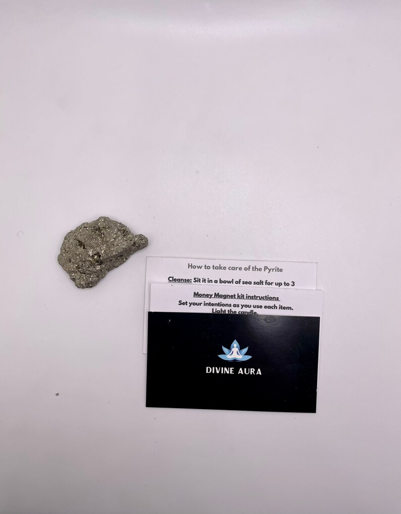 May include: A small, gray, metallic rock with a rough texture. A black card with white text reads "How to take care of the Pyrite Cleanse: Sit it in a bowl of sea salt for up to 3 Money Magnet kit instructions Set your intentions as you use each item. Light the candle. DIVINE AURA"
