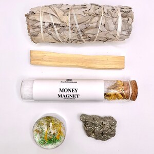May include: A white sage smudge stick, a palo santo stick, a glass vial with dried herbs labeled "MONEY MAGNET Spiritual Bath", a green and gold glitter candle, and a piece of pyrite.