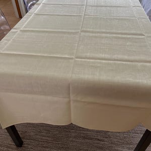 May include: A rectangular off-white tablecloth with a subtle woven texture and a scalloped edge. The fabric features a grid-like pattern of visible fold lines across the surface, covering the top of a dark-legged table in a dining space.
