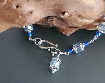 May include: A silver bracelet with a silver clasp and blue crystal beads. The bracelet is on a piece of driftwood.