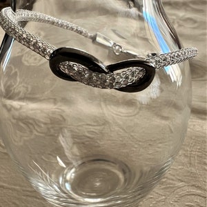 May include: A silver-toned bracelet with a black oval centerpiece rests on a clear glass vase. The bracelet features a woven chain design and a clasp closure. The vase is empty and sits on a patterned surface.