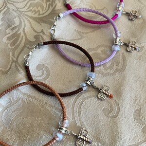 May include: Four braided cord bracelets with silver dragonfly charms and white glass beads. The bracelets are in shades of pink, purple, brown, and beige.