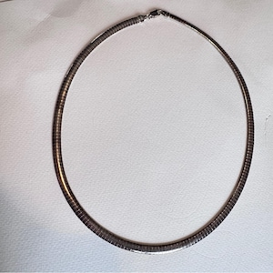May include: A silver-toned necklace with a flexible, ribbed design. The necklace has a smooth, rounded shape and a clasp closure. The jewelry is displayed on a white surface.