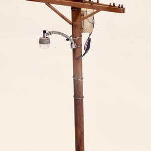 May include: A miniature wooden telephone pole with crossarms, insulators, and a small light fixture. The pole is brown with metal wire wrapping and a small base with faux grass. A small box is attached to the pole.