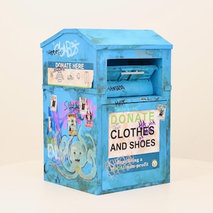 May include: A light blue donation bin for clothes and shoes, covered in graffiti and stickers. The bin has a slanted roof and a slot for donations. Text on the bin reads "DONATE CLOTHES AND SHOES" and "DONATE HERE".