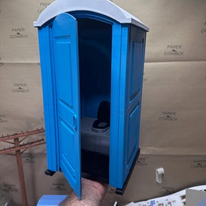 May include: A miniature blue portable toilet with a white roof and open door. The interior features a black toilet seat and a dark floor mat. The model is a novelty item.
