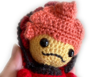 May include: A crocheted plush toy with a red body, black accents, and a pink and red crocheted hair. The toy has a yellow face with black eyes and a black mouth.