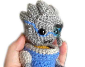 May include: A crocheted plush toy in the shape of a robot. The robot is gray with blue and yellow accents. It has a blue eye and a gray face.
