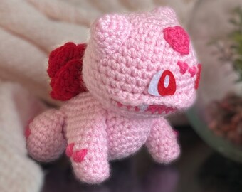 May include: Pink crocheted amigurumi toy with red heart details. The toy is shaped like a small creature with a large head and a small body.