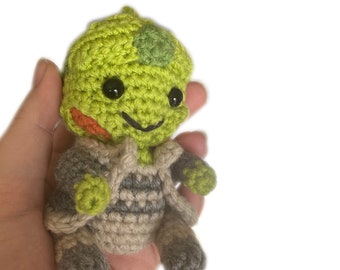 May include: A green crocheted plush toy with a gray crocheted outfit. The toy has black eyes and a smiling mouth.