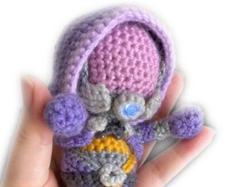 May include: A crocheted plush toy in the shape of a purple and gray alien with yellow accents. The toy has a large head with a purple hood and a blue eye.