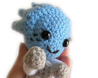 May include: A crocheted plush toy in the shape of a person with blue skin, white clothing, and gray arms and legs. The toy has black eyes and a smiling mouth.