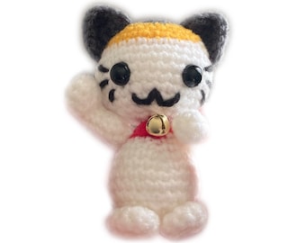 May include: White crocheted cat with black ears and a yellow stripe on its head. The cat is wearing a red collar with a gold bell.