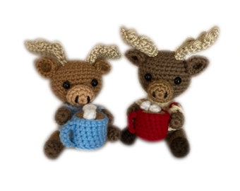 May include: Two brown crocheted moose holding mugs of hot chocolate with marshmallows. The moose on the left is holding a blue mug and the moose on the right is holding a red mug.