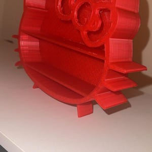 Pode incluir: Uma prateleira vermelha impressa em 3D em forma da cabeça de Hello Kitty. A prateleira tem vários níveis horizontais para armazenamento e um detalhe de laço na parte superior. O design é de cor vermelha vibrante e assenta numa superfície branca.