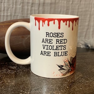May include: White ceramic mug with a red dripping rim. The mug has a black graphic that says "Roses are red, Violets are blue". The graphic also includes a stylized red rose with green leaves.