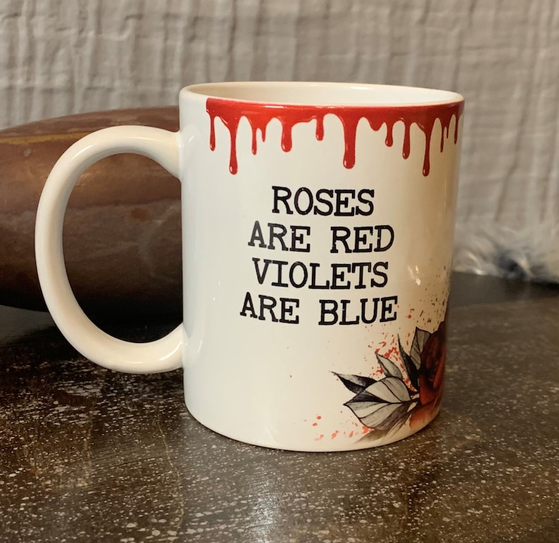 May include: White ceramic mug with a red dripping rim. The mug has a black graphic that says "Roses are red, Violets are blue". The graphic also includes a stylized red rose with green leaves.