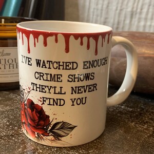 May include: White ceramic mug with red dripping paint around the rim. The mug has a black graphic of a rose with red accents and the text "I've watched enough crime shows they'll never find you."