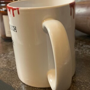 May include: White ceramic mug with a red dripping paint design around the rim. The mug has a black inscription that reads "You're Enough".