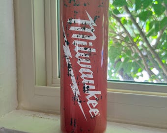 May include: A red tumbler with the word "Milwaukee" in white, featuring a distressed design. The tumbler is cylindrical and appears to be made of metal. The background is a window with green foliage.