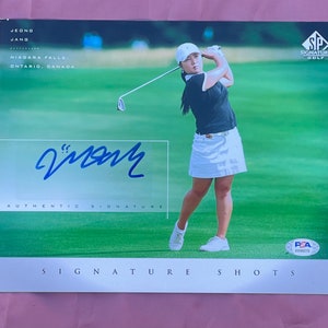 Puede incluir: Una tarjeta de golf blanca y negra con una mujer con una camisa negra y una falda blanca balanceando un palo de golf. La tarjeta tiene el texto "SIGNATURE GOLF" y "JEONG JANG" en las esquinas superior derecha e izquierda, respectivamente. La tarjeta también tiene el texto "NIAGARA FALLS, ONTARIO, CANADA" y "SIGNATURE SHOTS" en las esquinas inferior izquierda y derecha, respectivamente. La tarjeta tiene una firma azul que dice "Jjang" y el texto "AUTHENTIC SIGNATURE" debajo. La tarjeta también tiene una pegatina PSA clasificada en la esquina inferior derecha.