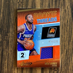 Op de afbeelding: Een basketbal trading card met Mikal Bridges van de Phoenix Suns. De kaart is uit de Rookie Threads-serie en is genummerd 079/149. De kaart toont een foto van Bridges in zijn Suns-uniform, een stukje van zijn jersey en de tekst "Rookie Threads" en "Absolute Memorabilia".